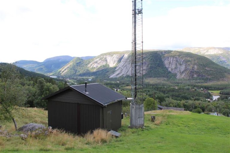 Basestasjon med teknisk bygg og høy mobilmast plassert på en gresskledd høyde. I bakgrunnen sees en dal med skog, elv og bratte fjell.